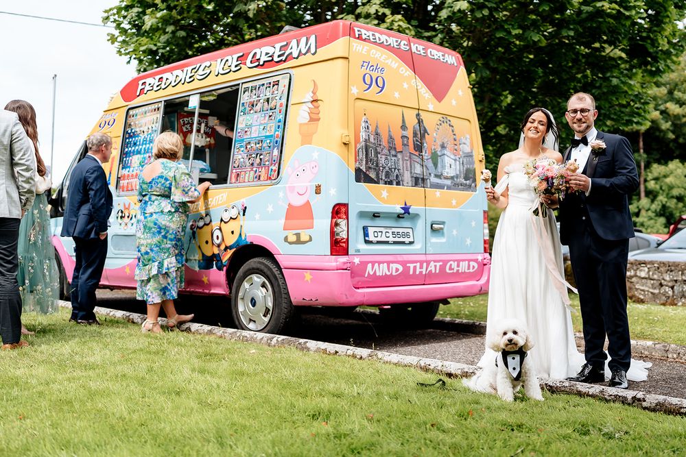 Colorful ice cream truck parked on grass serves wedding guests while newlyweds stand nearby with their dog.