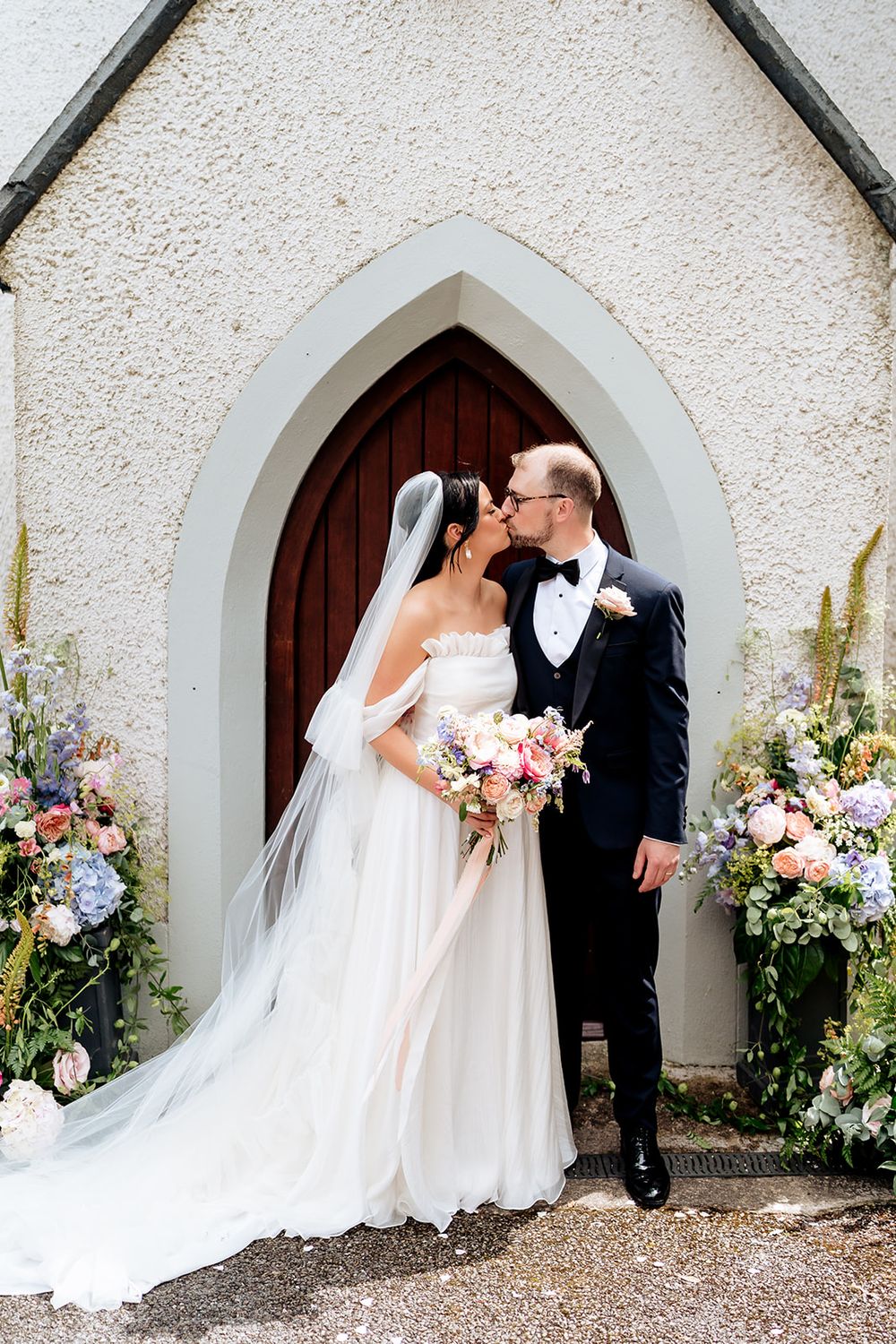 Newlywed couple kissing in front of church doorway surrounded by pink and purple floral arrangements.