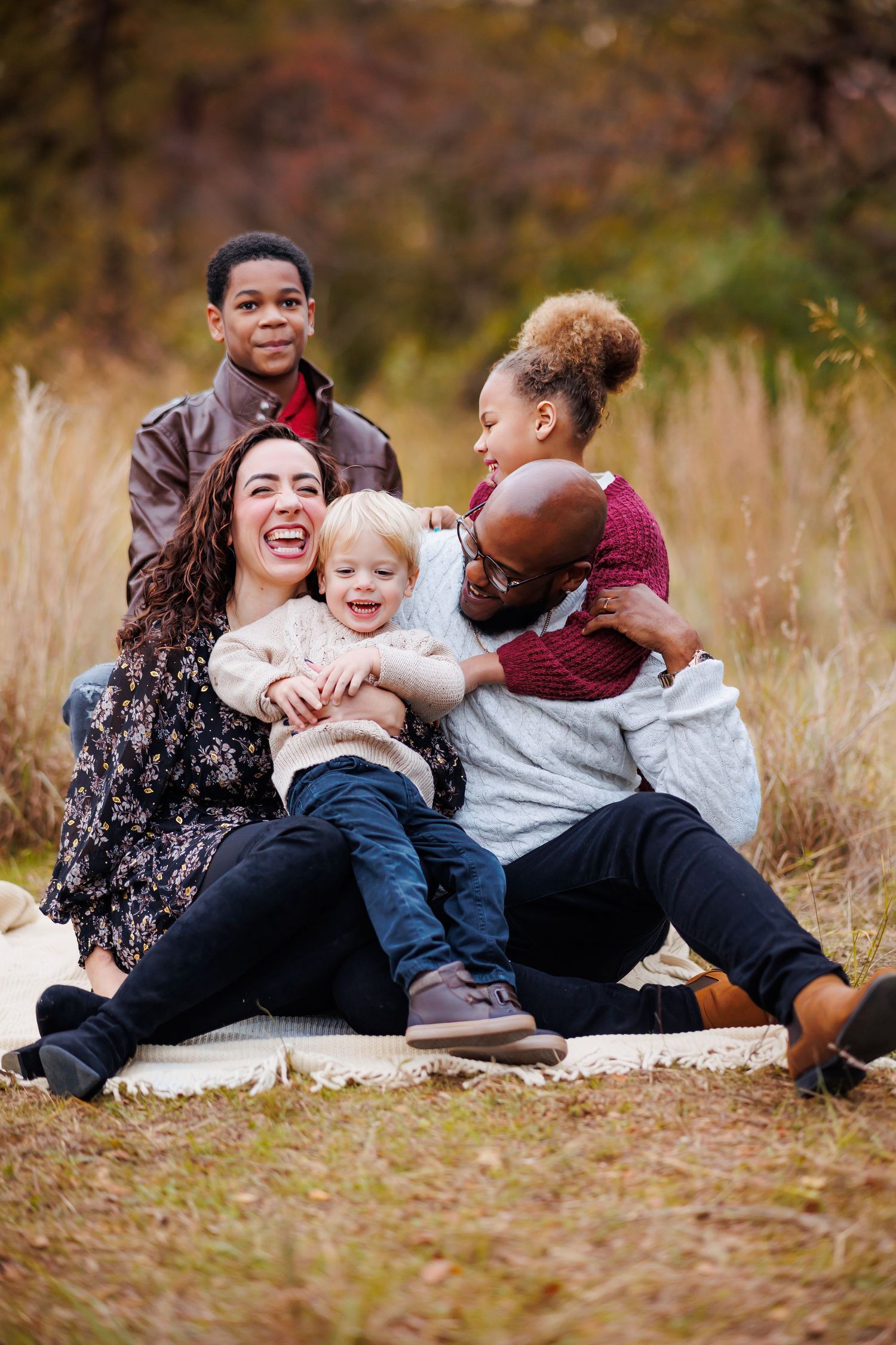 A family sits together laughing while posing for a portrait in a field of tall grass during autumn wearing mixed blues and burgundy outfits