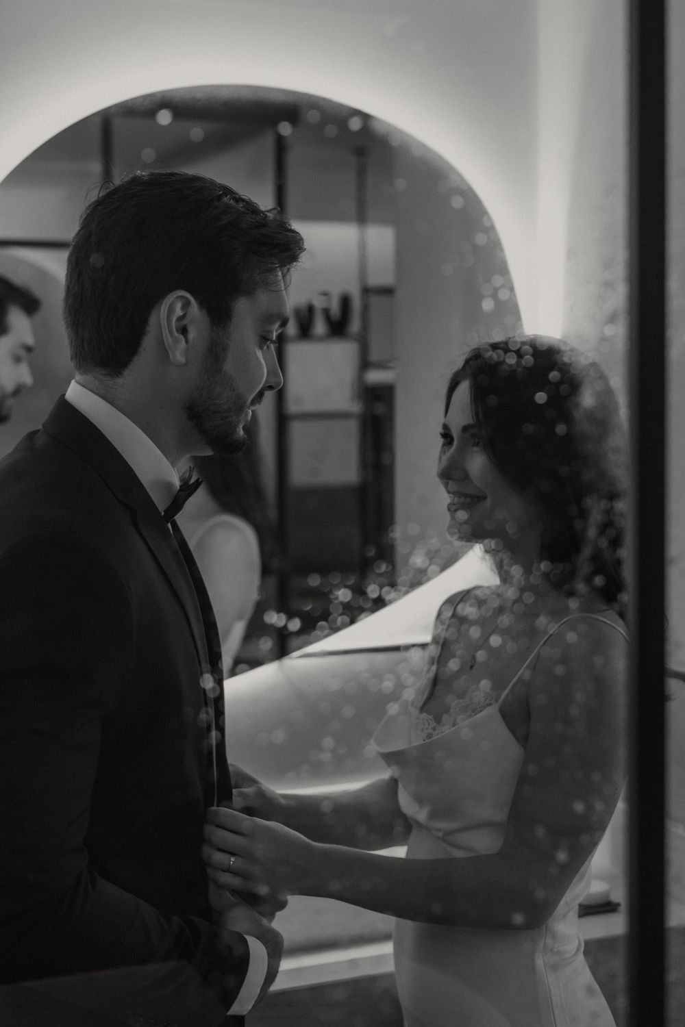 A romantic black and white wedding photo of a couple gazing at each other under an archway with soft lighting.