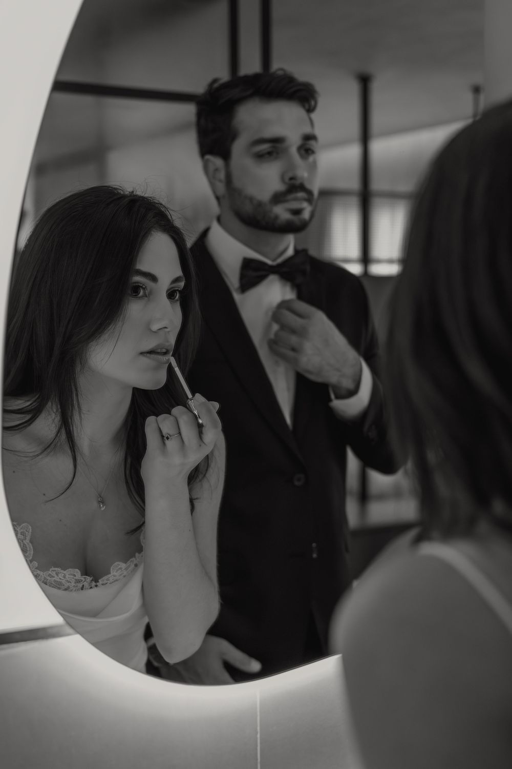 Black and white photo of a couple adjusting their formal wear while looking in a mirror during a wedding preparation.