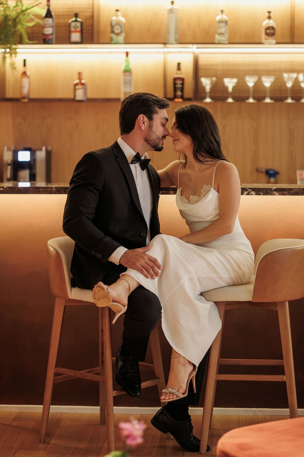 A romantic couple shares an intimate moment at an elegant bar with backlit bottles and cocktail glasses on display.