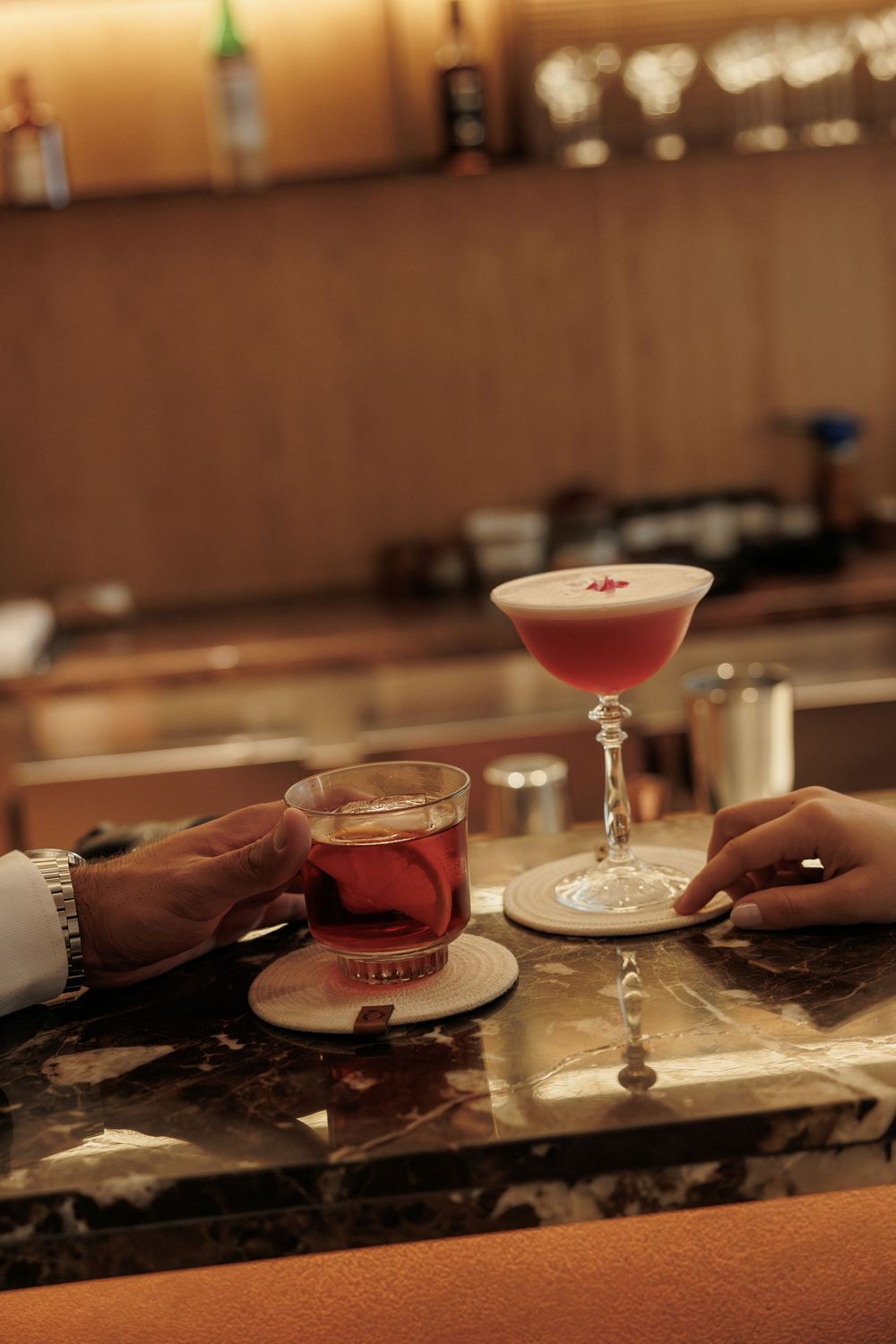 Close-up shot of cocktail glasses being placed on a dark marble bar counter in warm lighting.