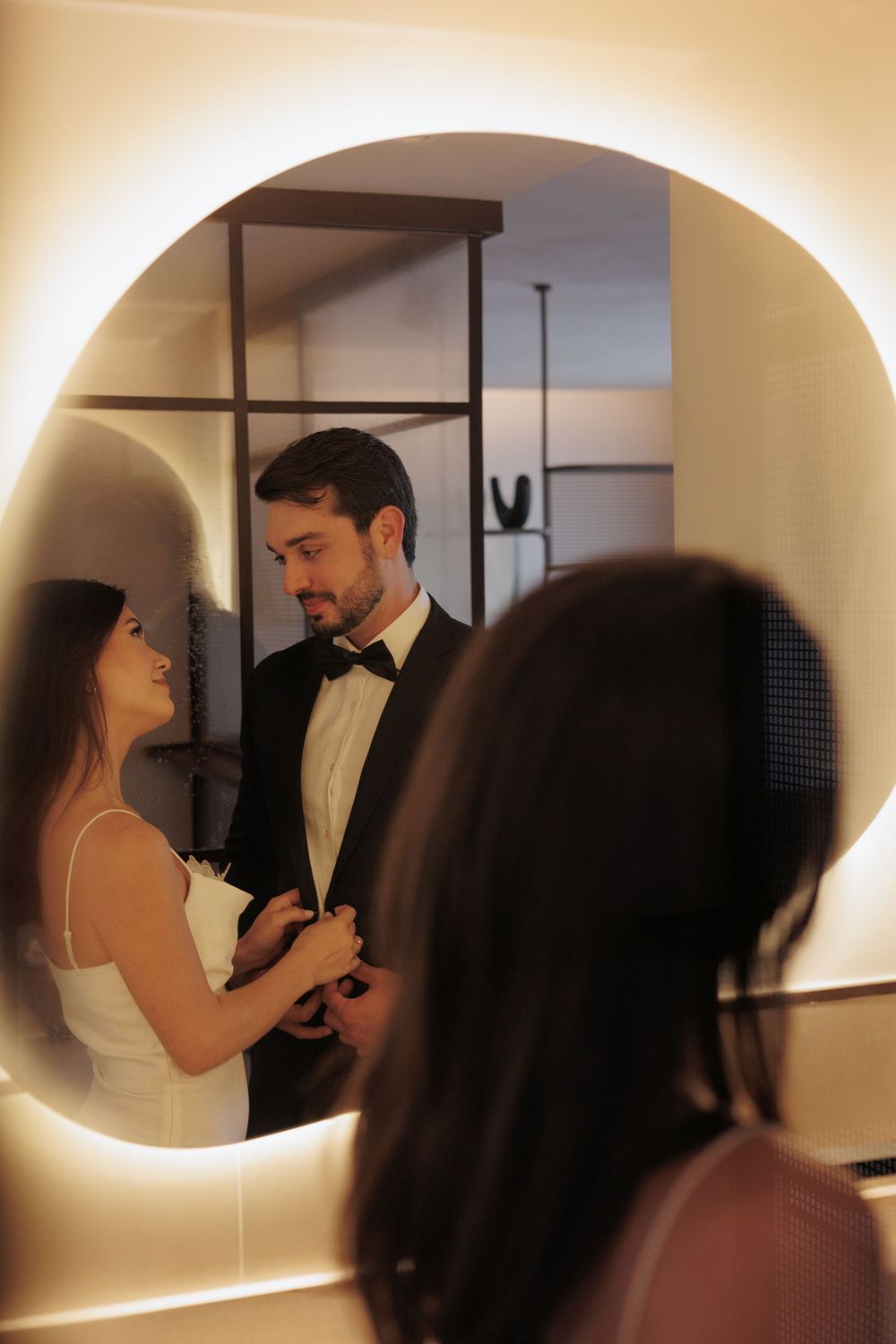 A couple shares an intimate moment while getting ready in formal wear as they're reflected in a mirror with soft lighting.