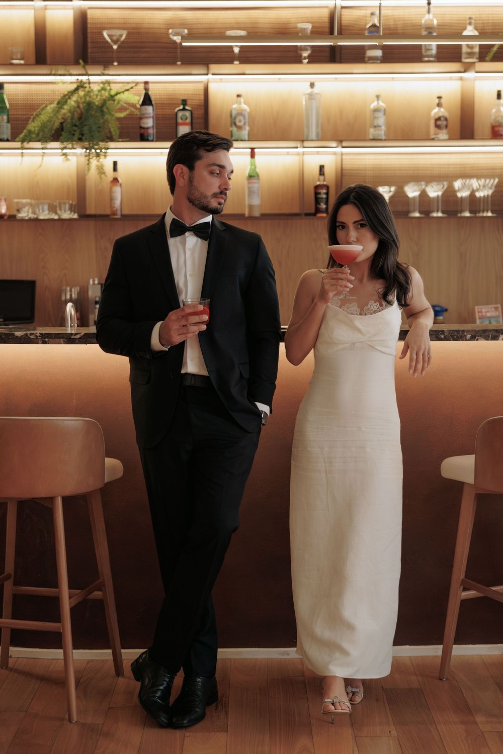 Elegantly dressed couple enjoying cocktails at an upscale bar with illuminated shelves of spirits behind them.