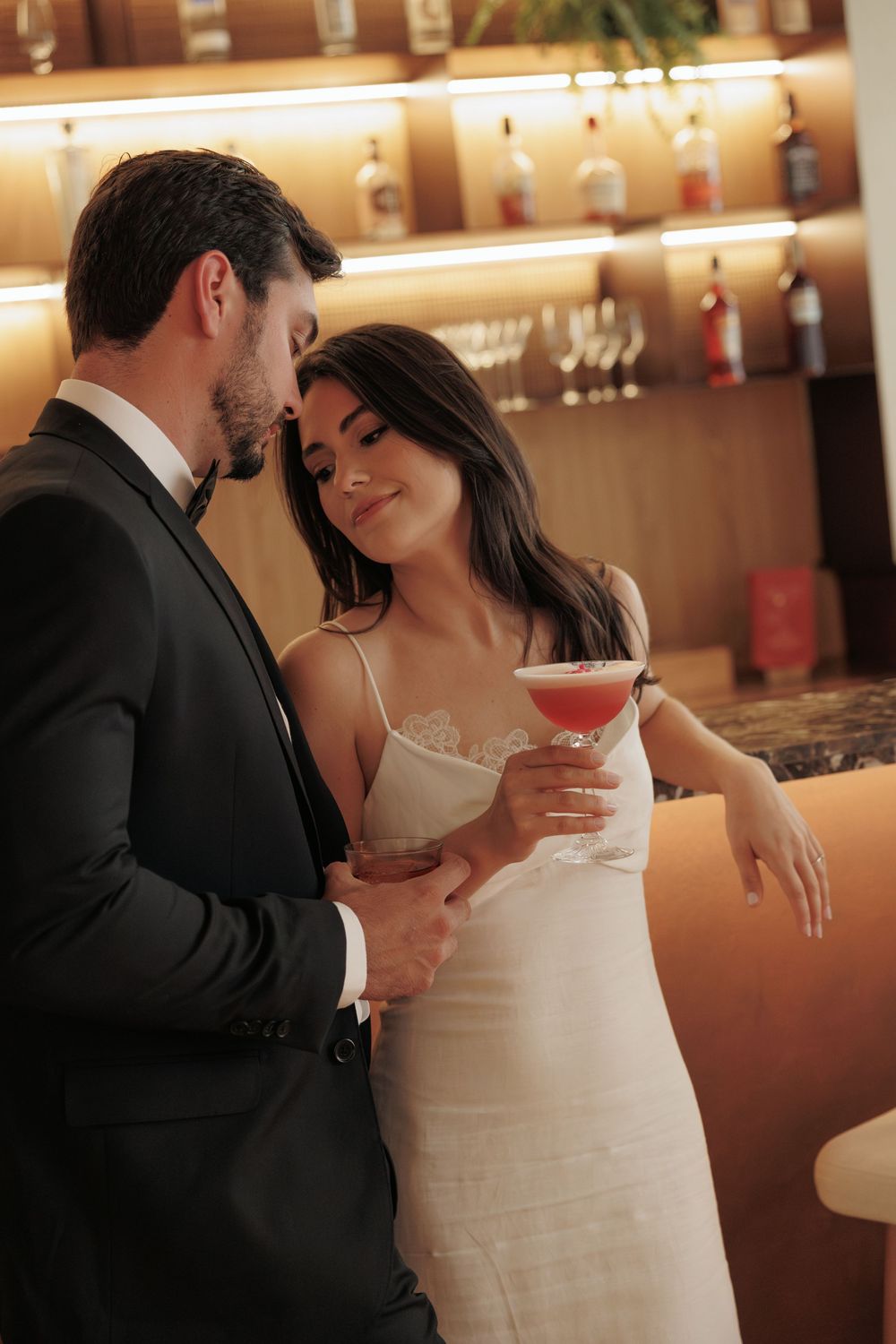 Couple in formal attire share intimate moment at upscale bar while holding cocktails.
