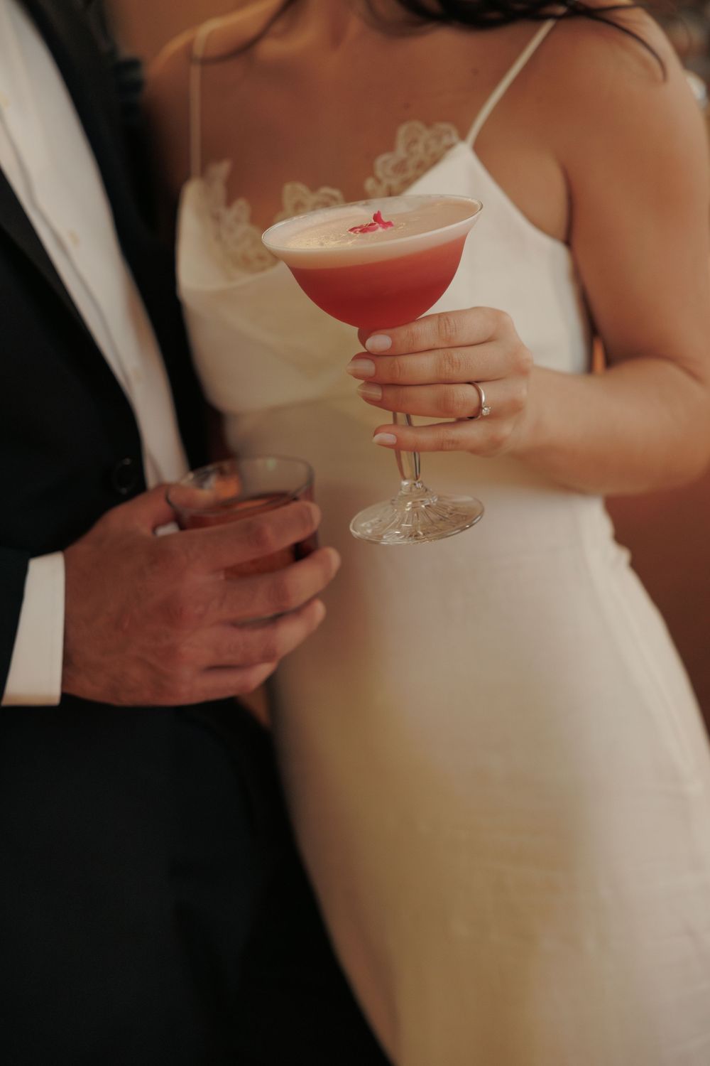 Close-up of someone in a white wedding dress holding a red cocktail while standing next to a person in a dark suit.