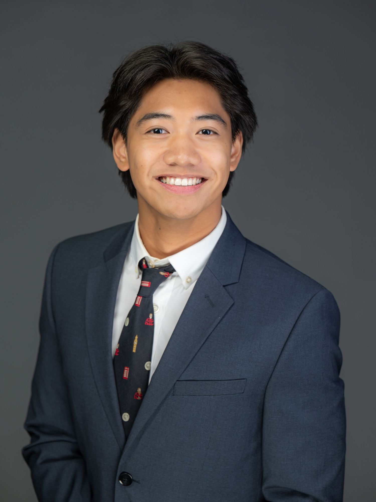 Professional headshot of someone in a navy suit and patterned tie smiling warmly against a gray background.