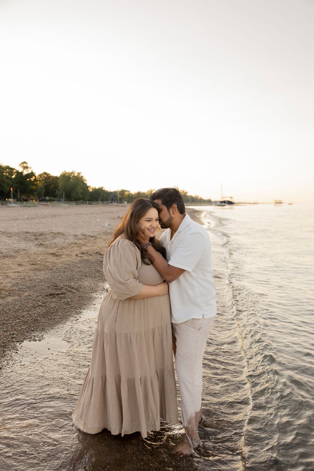 A pregnant couple shares loving moments as the sun sets on a scenic beach with gentle waves lapping at the shore.