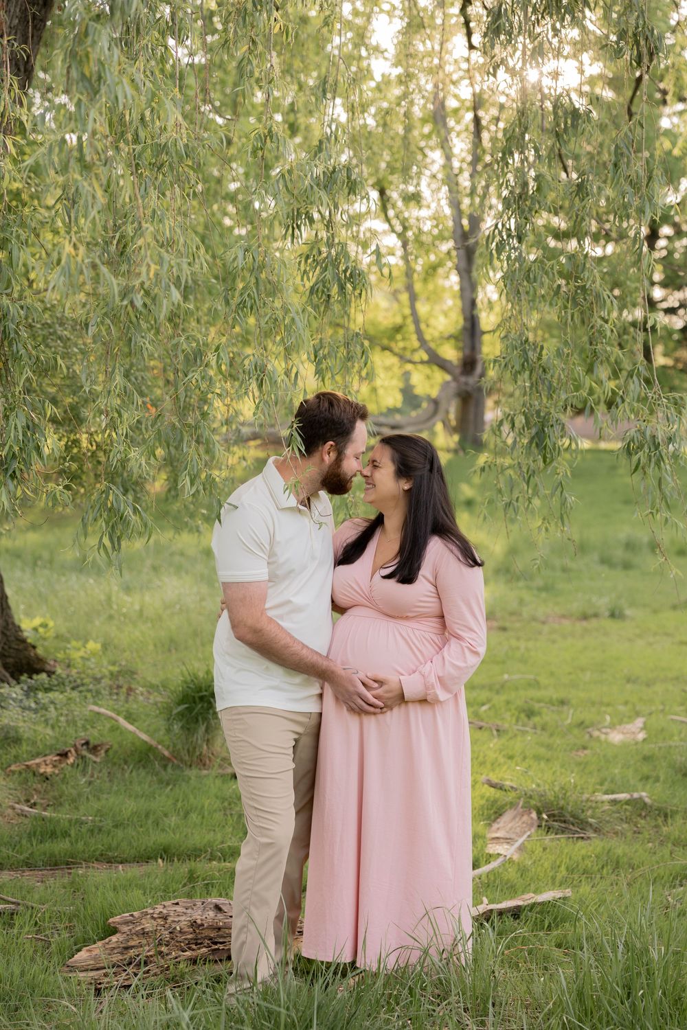 A romantic maternity photo shows a couple embracing under a weeping willow tree in Lake Orion michigan