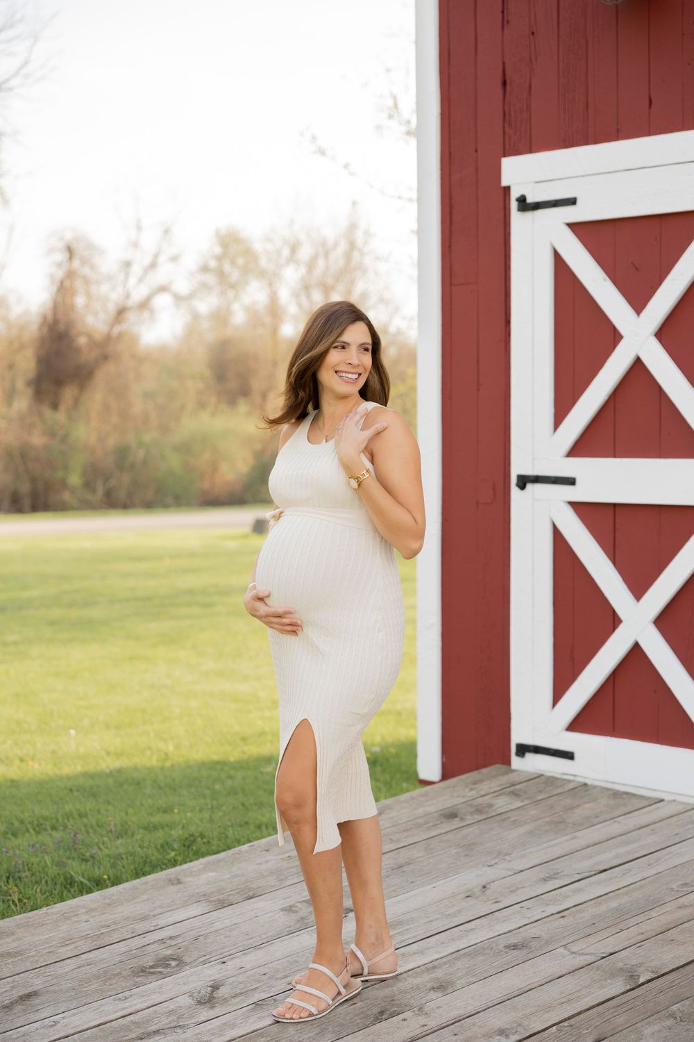 Expectant mother in white dress poses on wooden deck in front of red barn door on sunny day in Oakland country Rochester hills michigan