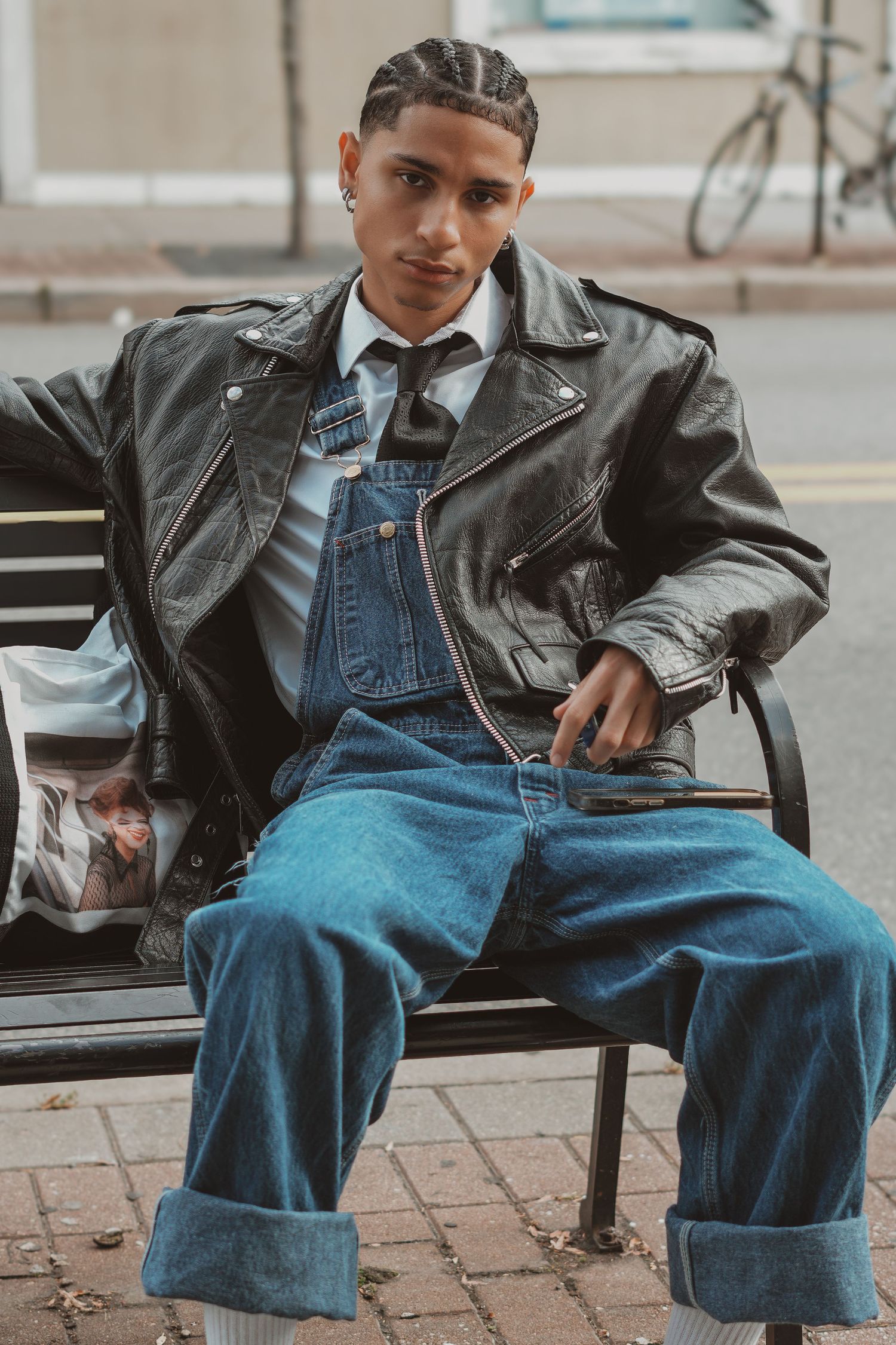 Casual model in denim overalls and leather jacket seated on bench.