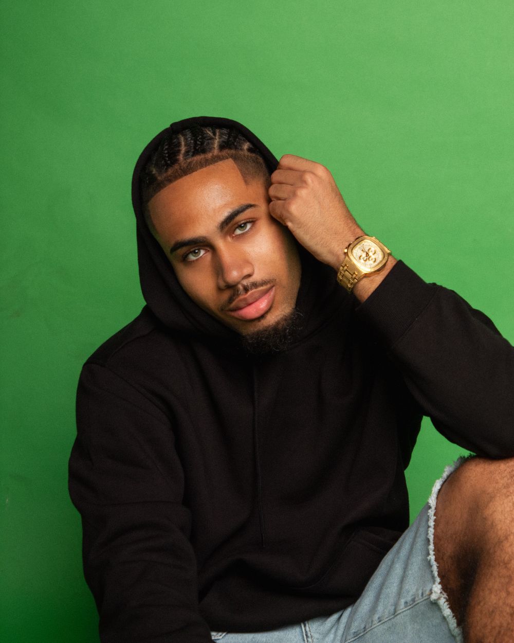 Portrait against green backdrop featuring person in black hoodie wearing gold watch.