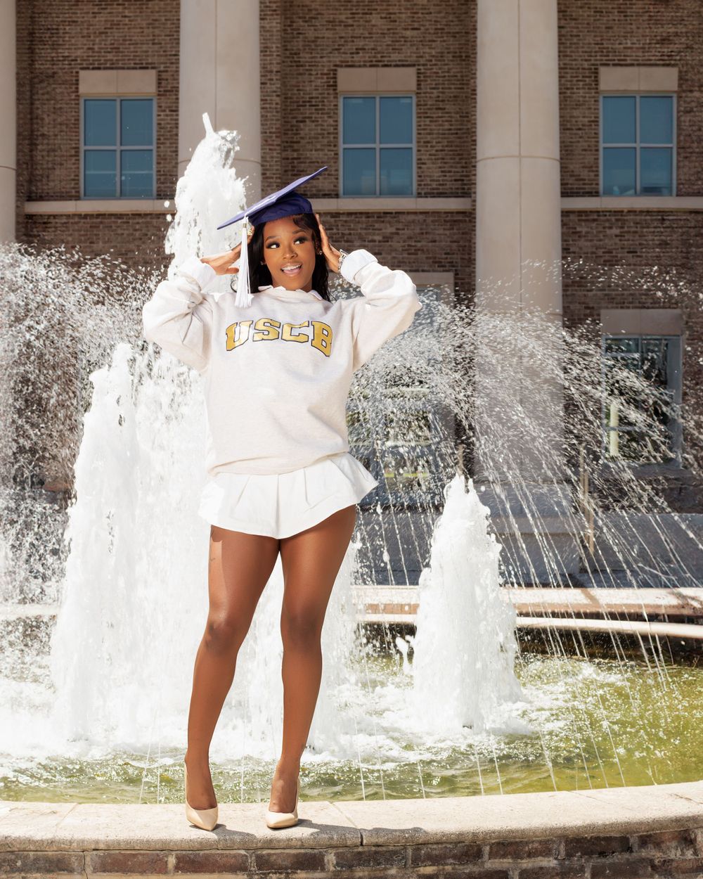 Graduate celebrating with water fountain backdrop wearing a white university sweatshirt and blue graduation cap.