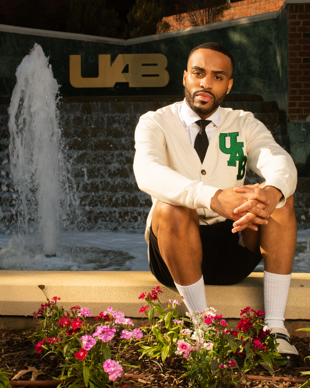 Graduate in white suit posing by UAB fountain with flowers in foreground.