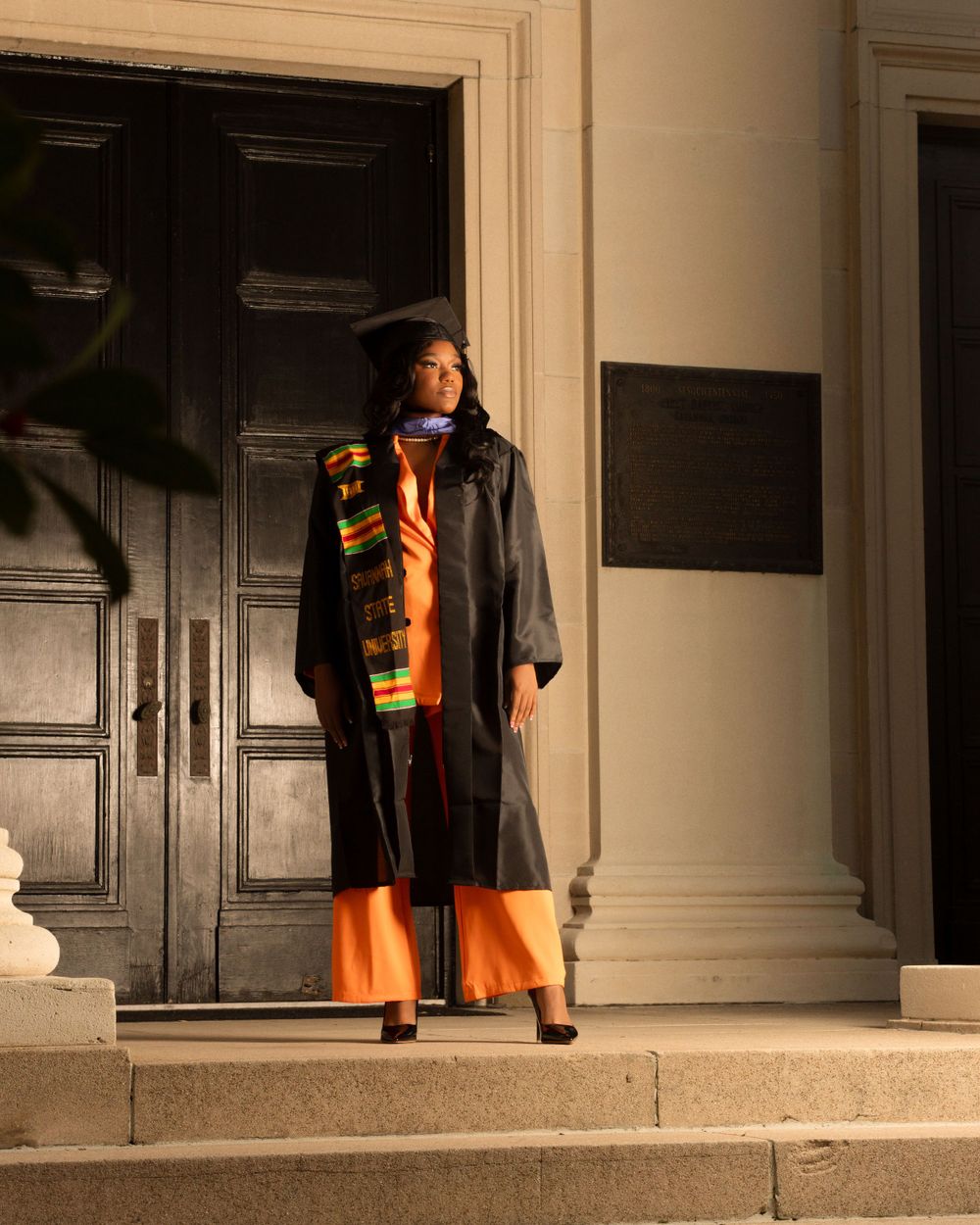 Graduate in academic regalia and colorful stole poses on steps by dark wooden doors.