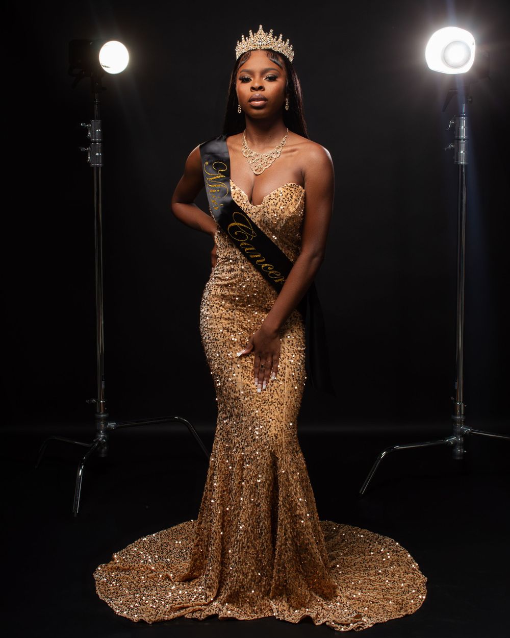 Glamorous gold sequined evening gown with a crown and sash photographed against a dark background with studio lights.