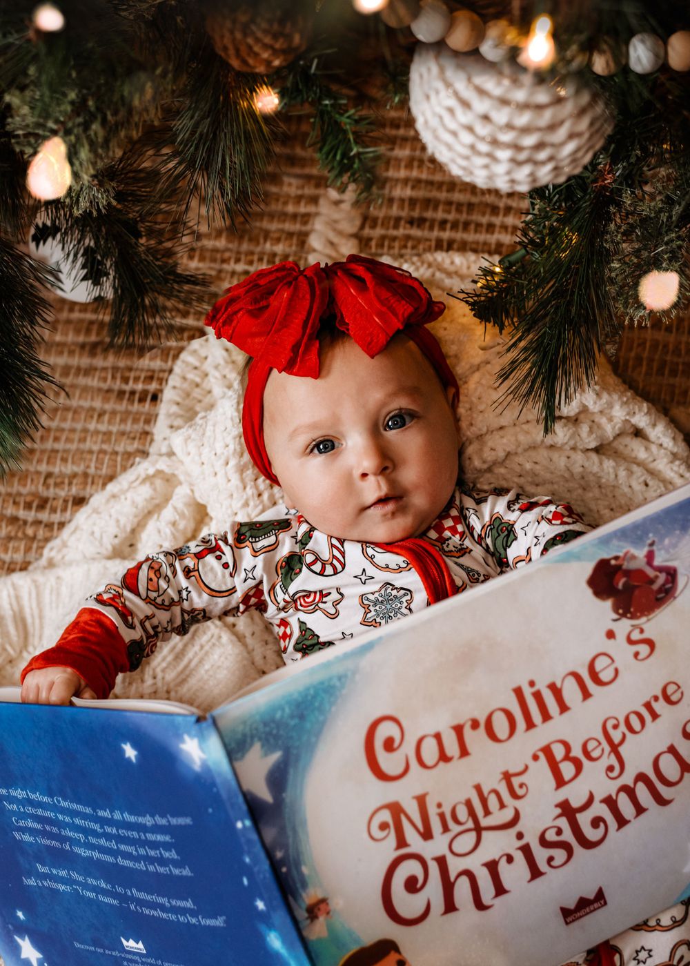 Baby in holiday outfit with red bow reads Christmas book.