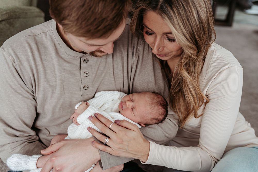 New parents in neutral colored sweaters tenderly hold their newborn baby.