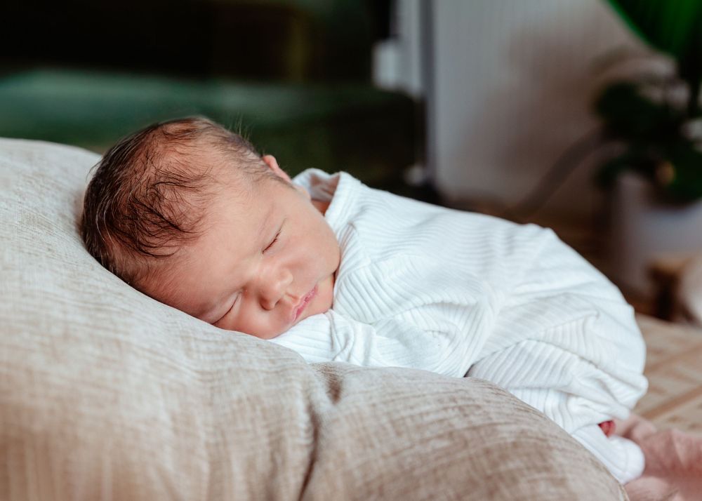A peaceful sleeping newborn in a white sweater rests on a textured beige pillow.