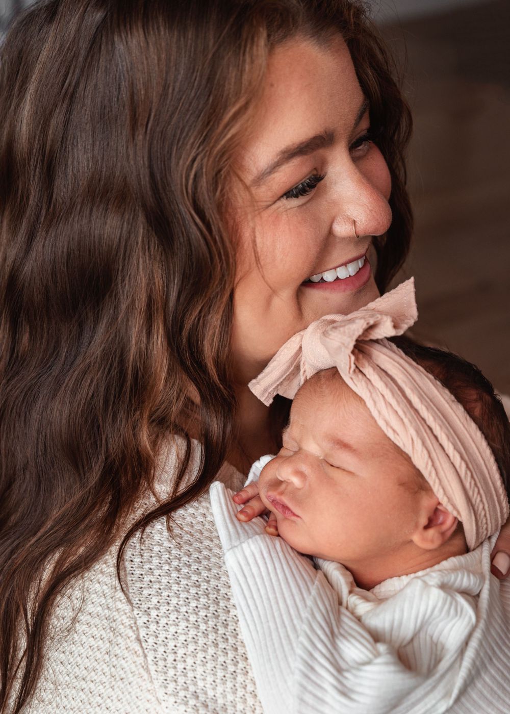A mother holding her sleeping newborn baby with a pink bow headband, wearing a white knit sweater.