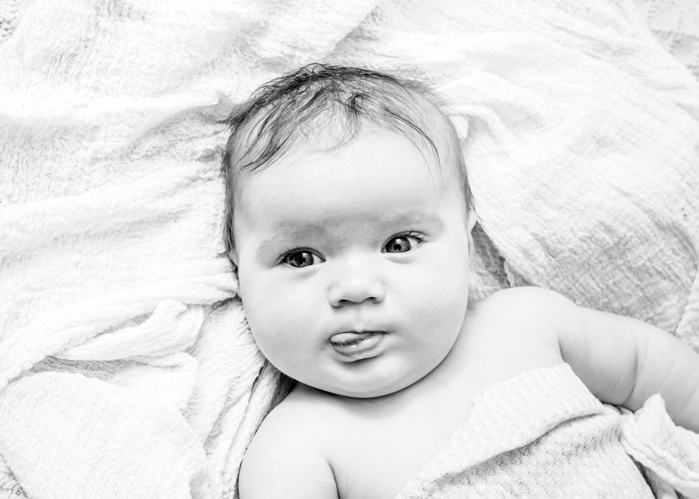 Black and white portrait of a smiling baby with chubby cheeks laying on white fabric.