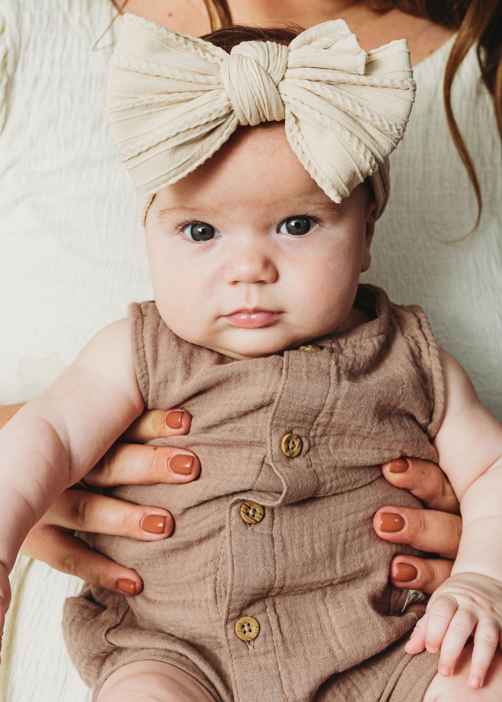 Baby wearing a cream bow headband and brown linen romper with buttons, with adult hands reaching in.