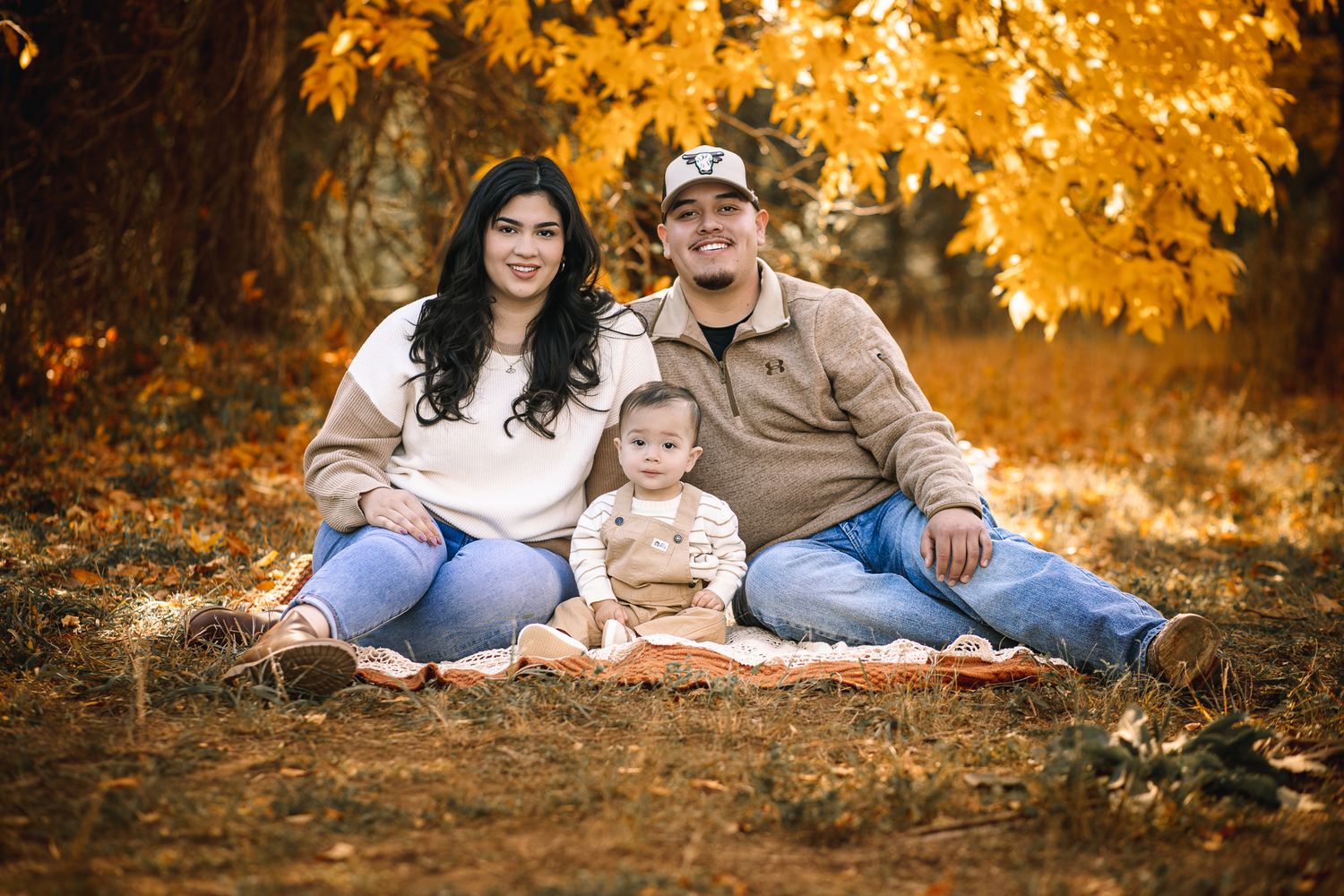 fall family photoshoot of a family of 3 sitting on the ground ith their baby. cripsy yellow and red leaves. sunset photoshoot.