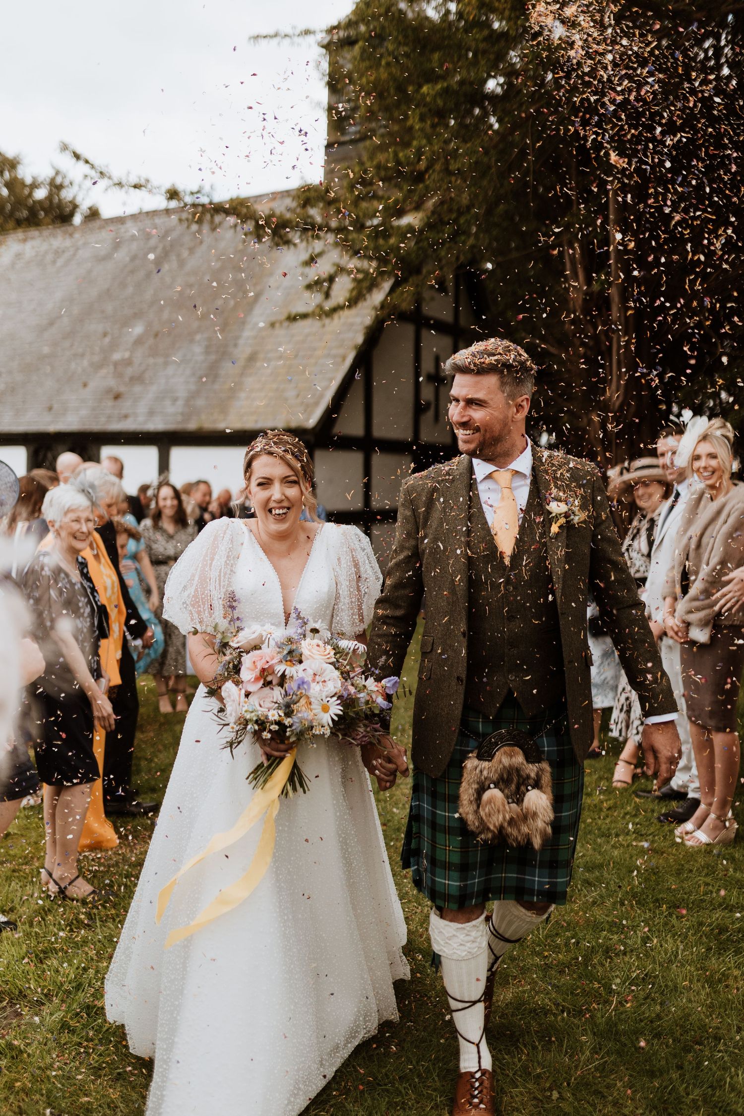 A newly married couple joyfully walks down a confetti-filled path at their Scottish themed outdoor wedding celebration at Quaint Country.