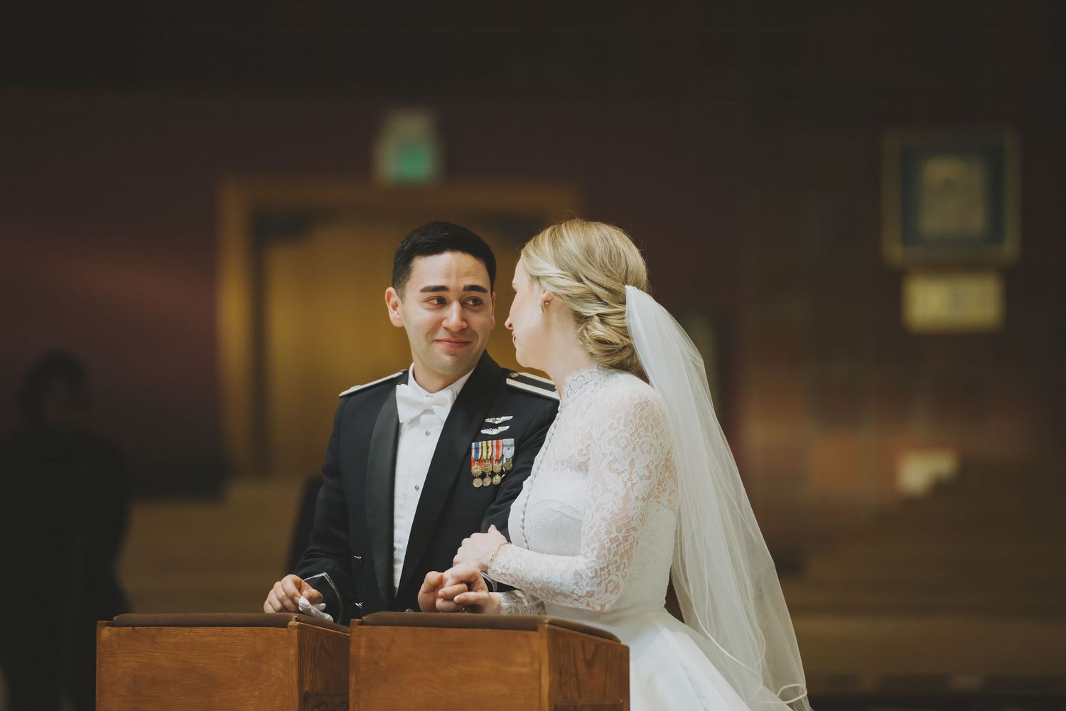 A bride and groom kneel together at a wooden podium during their indoor wedding ceremony.
