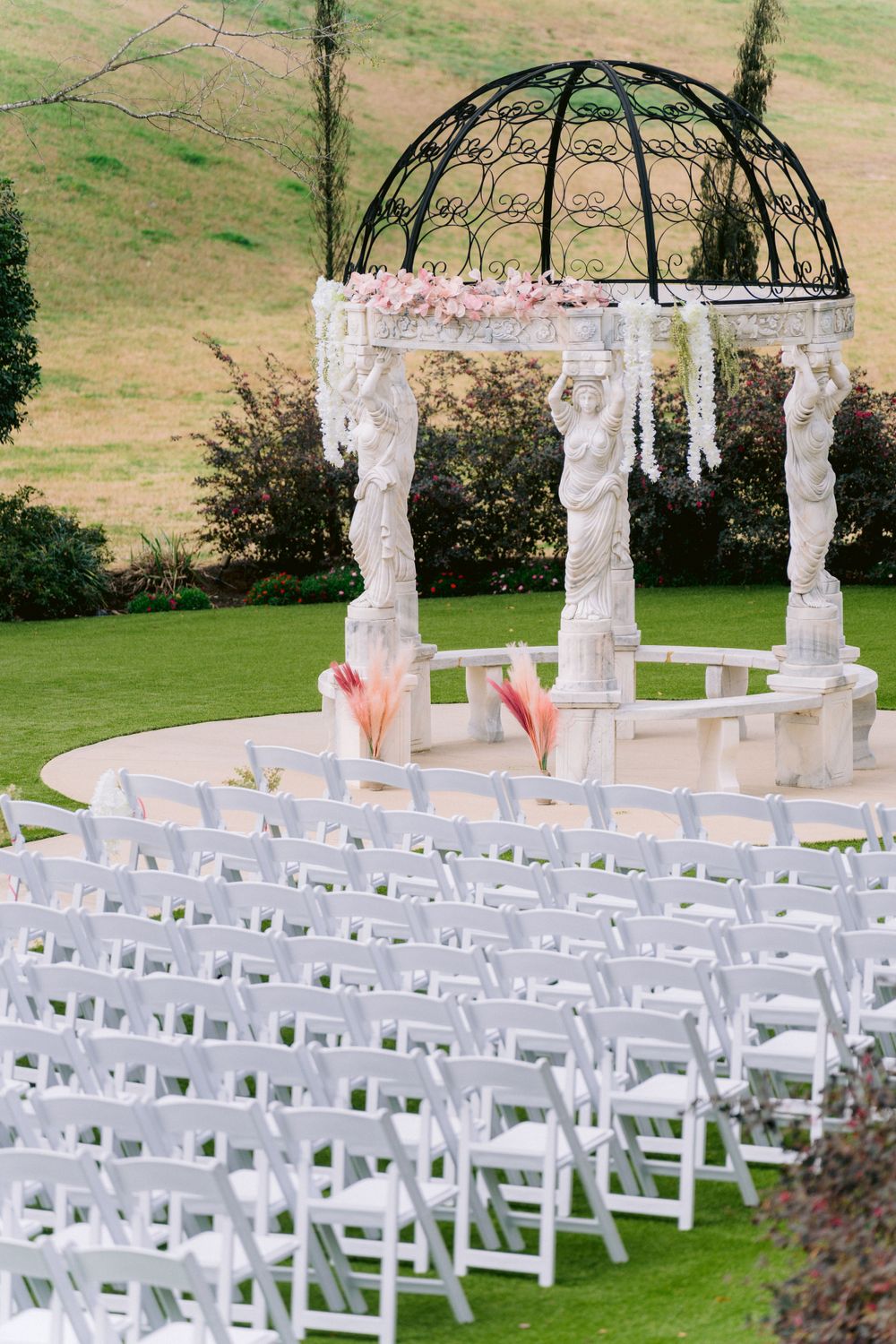 Outdoor wedding ceremony setup with white chairs facing an ornate gazebo decorated with pink flowers.