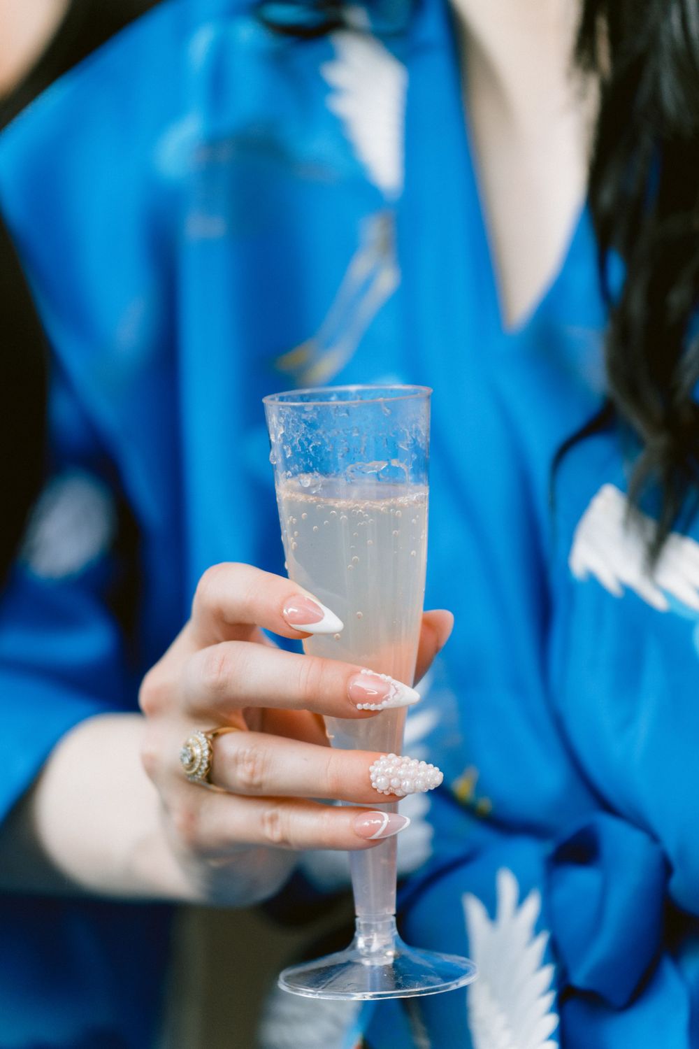 Champagne glass held by manicured hand wearing sparkly ring and blue silk robe.