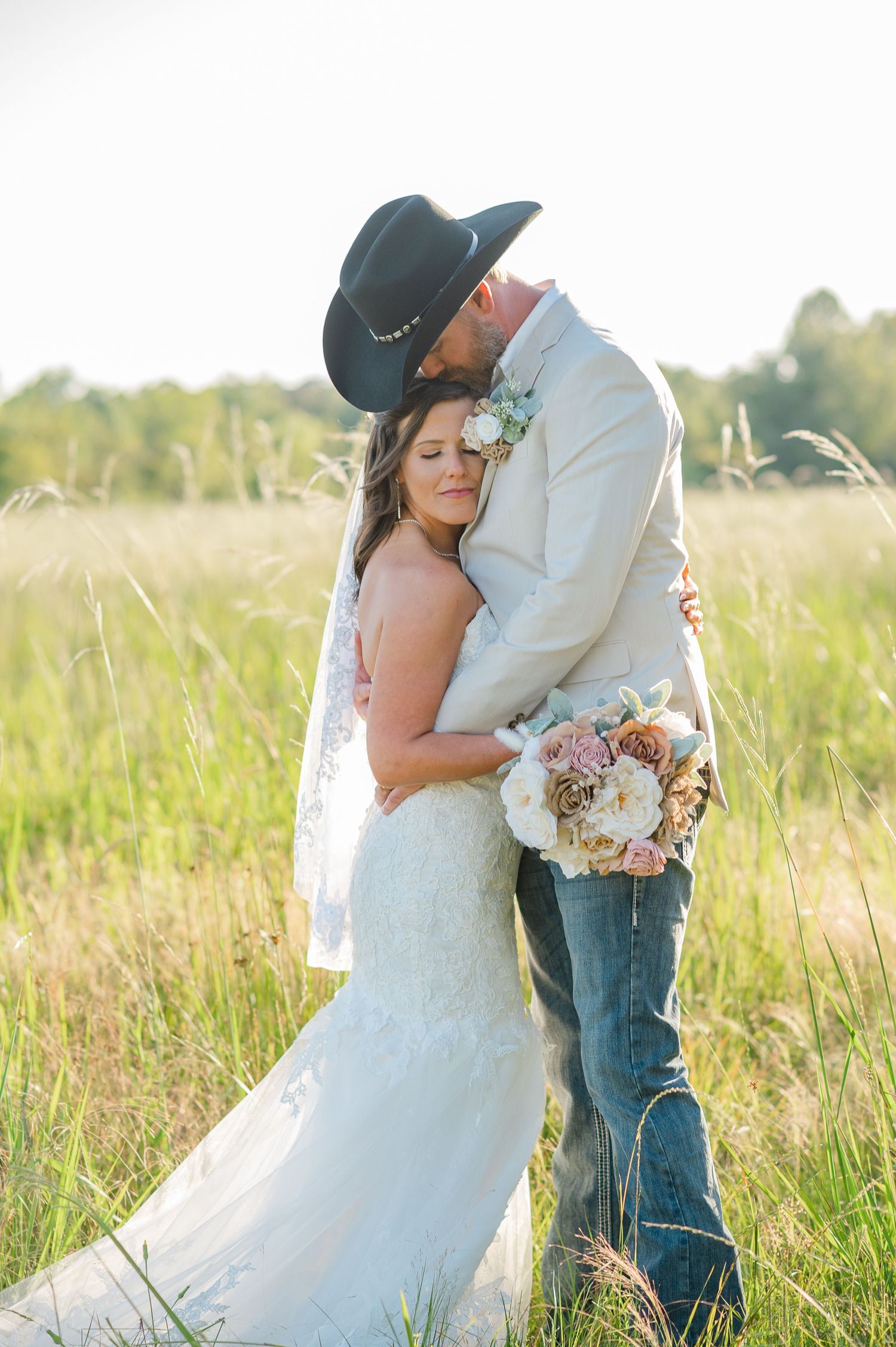 Romantic wedding portrait in golden field with bride holding bouquet and groom in western attire.