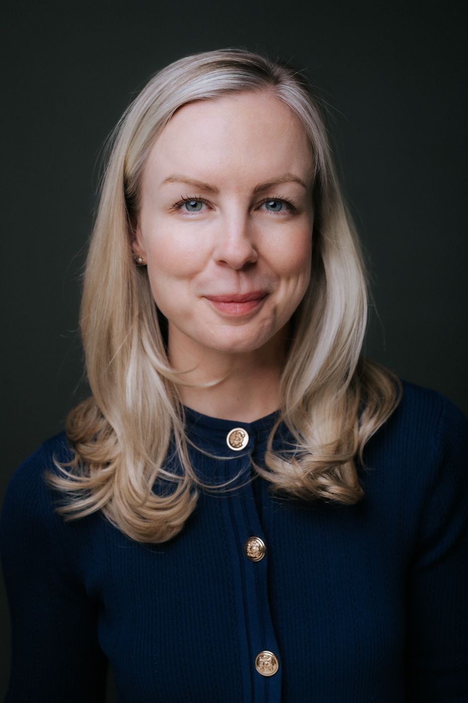 She wears a navy blue sweater adorned with gold buttons, exuding elegance and confidence in this well-lit, professional portrait.