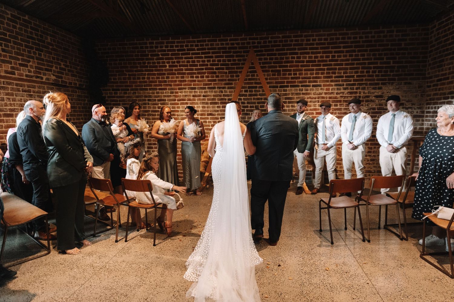 Wedding ceremony in a rustic brick venue with wooden chairs arranged in rows.