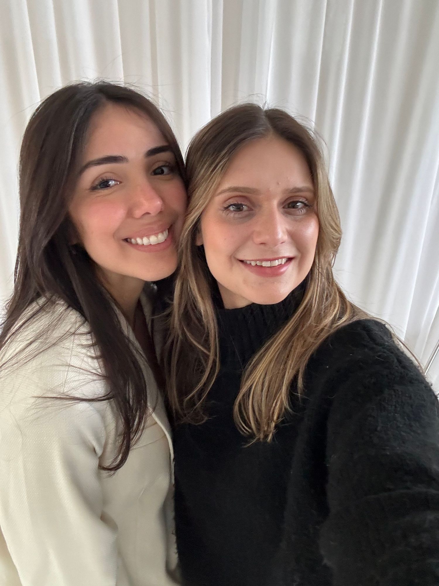 Two friends in winter sweaters taking a cozy selfie against white curtains.