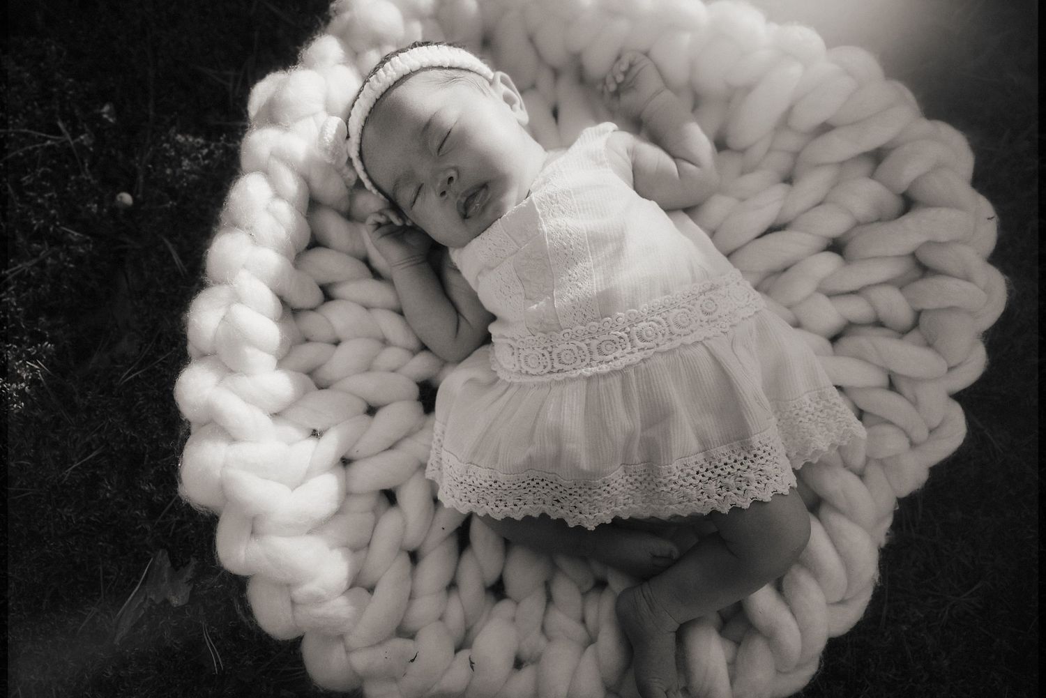 Sleeping baby wrapped in white lace dress lying on chunky knit blanket in black and white photograph.
