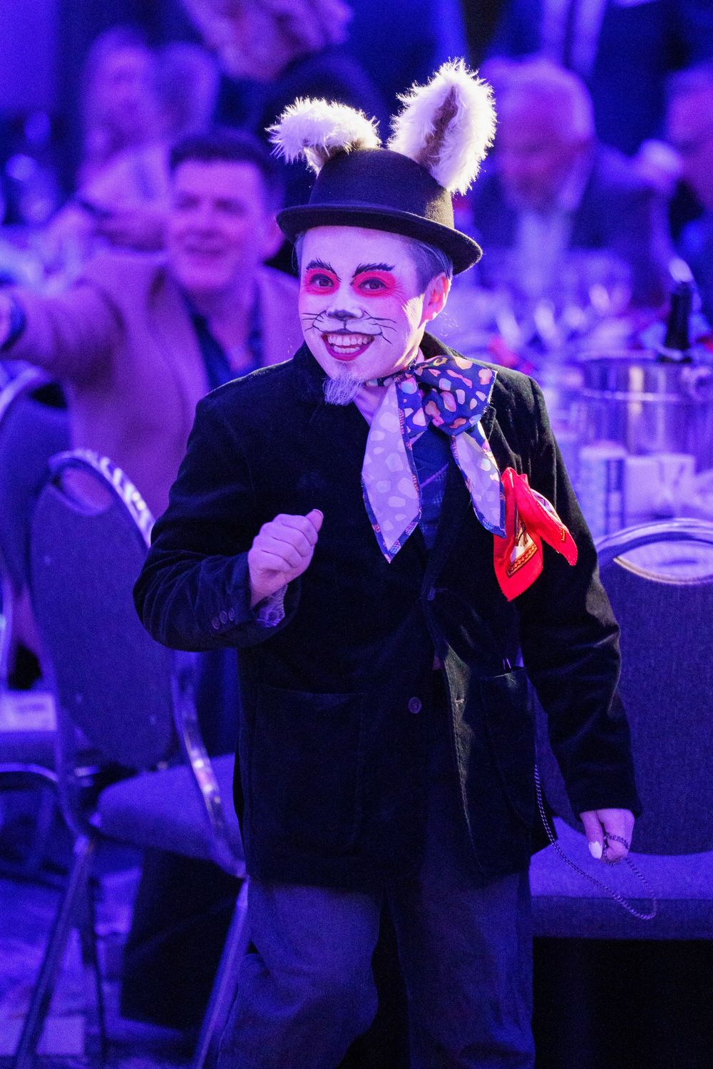 Performer in black suit with bunny ears and white face paint dances under purple stage lighting at an event.