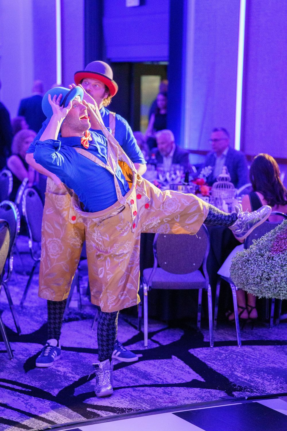 A performer in bright blue shirt and gold pants dances on stage under purple lighting at an indoor event.