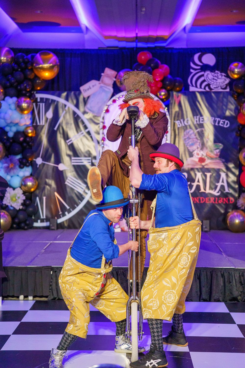 Three performers in matching blue and gold outfits execute an acrobatic routine on a checkered stage with colorful decor.