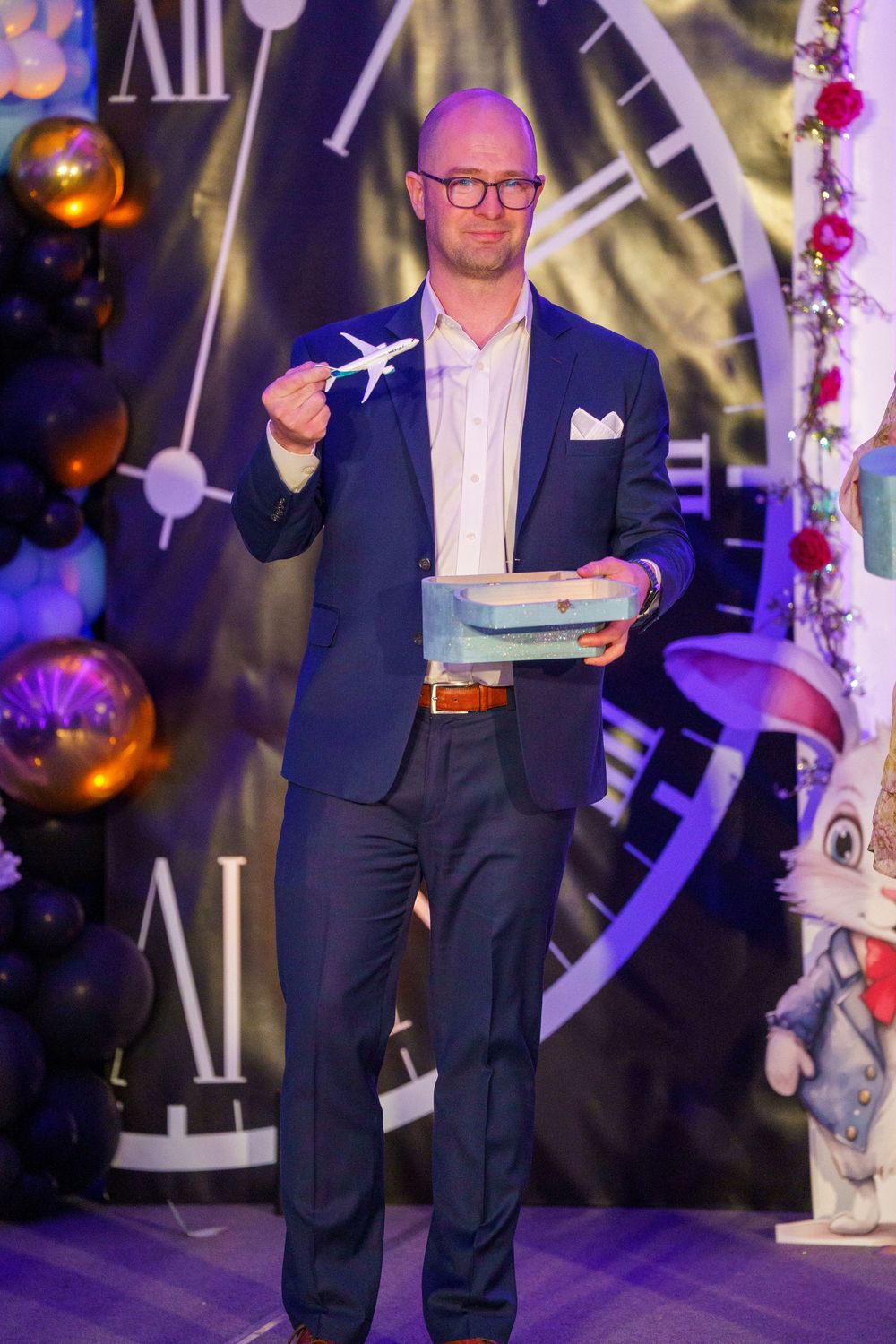 A person in a navy suit and glasses stands in front of a large decorative clock with holiday decorations and purple lighting.