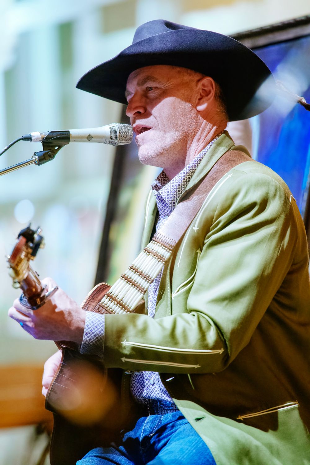 A country music performer in a cowboy hat and green jacket sings into a microphone while playing guitar on stage.