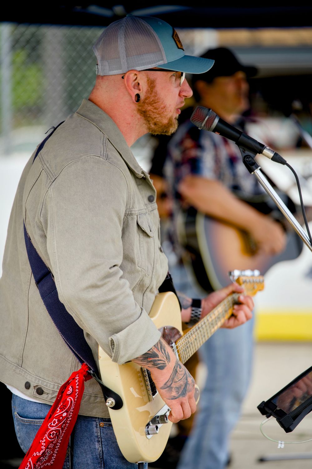 Musician performing on stage with a guitar wearing a light colored jacket and baseball cap.