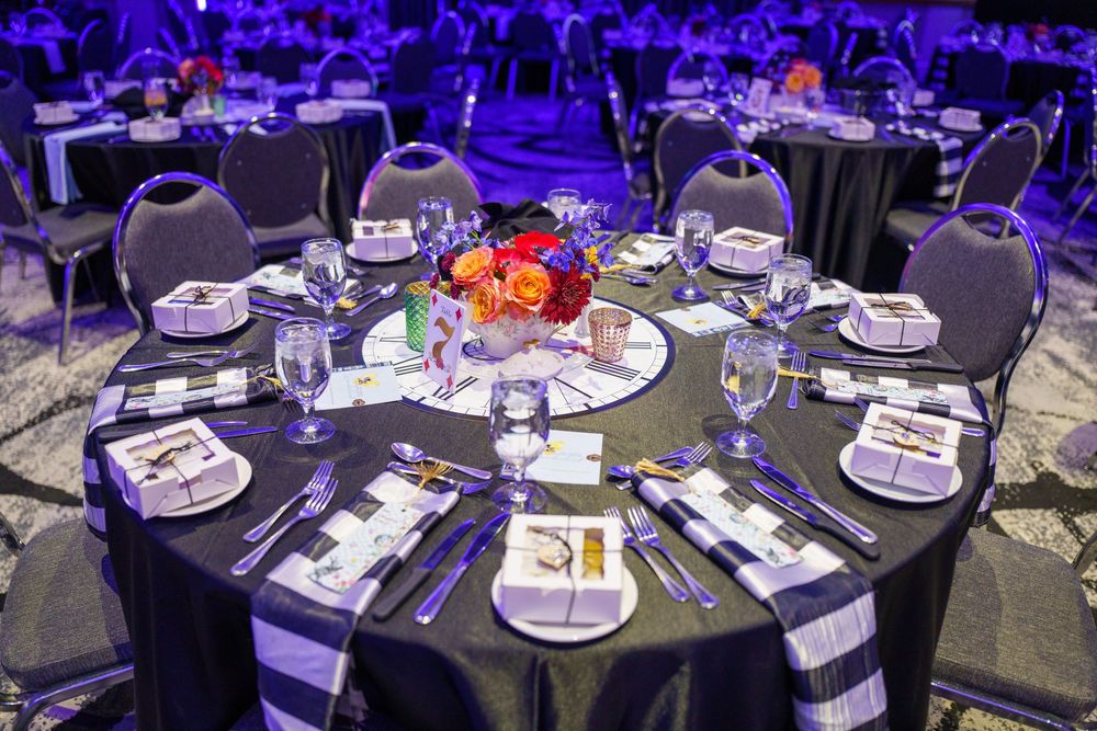 Elegant round table setting at formal event with dark linens, floral centerpiece and purple ambient lighting in ballroom.