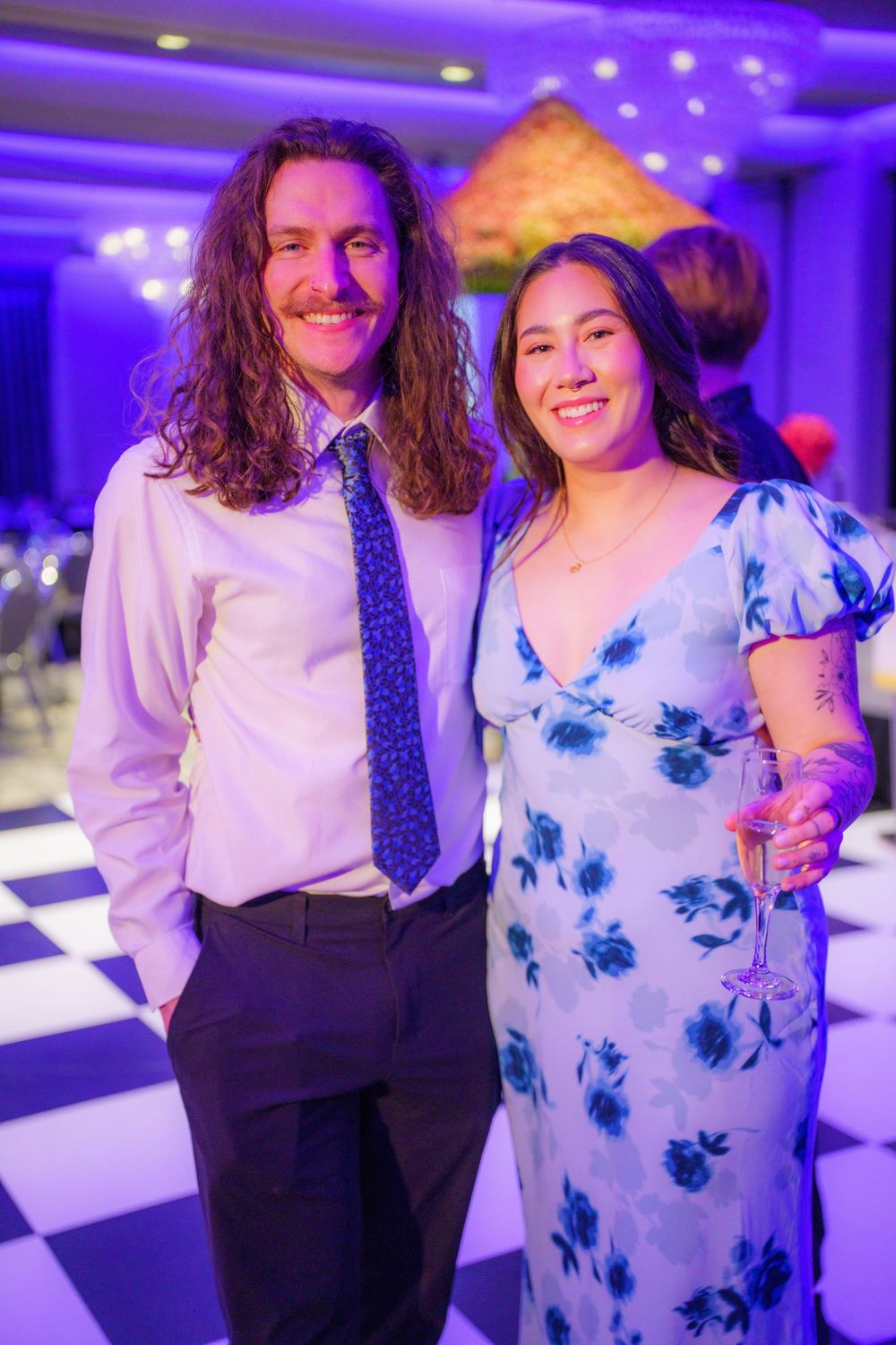 Two people pose at a formal event with a checkered dance floor and purple lighting, one wearing blue floral dress and one in tie.