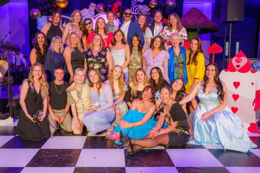 Large group of performers in colorful costumes posing together on a black and white checkered dance floor at an event.