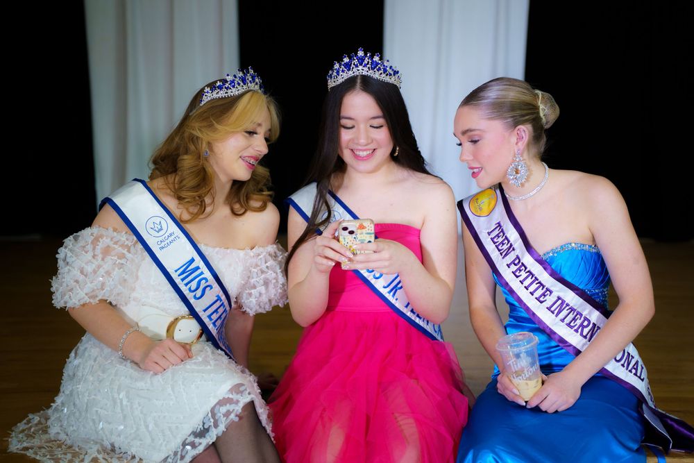 Three pageant winners wearing crowns and sashes share a moment looking at a phone together during an event.