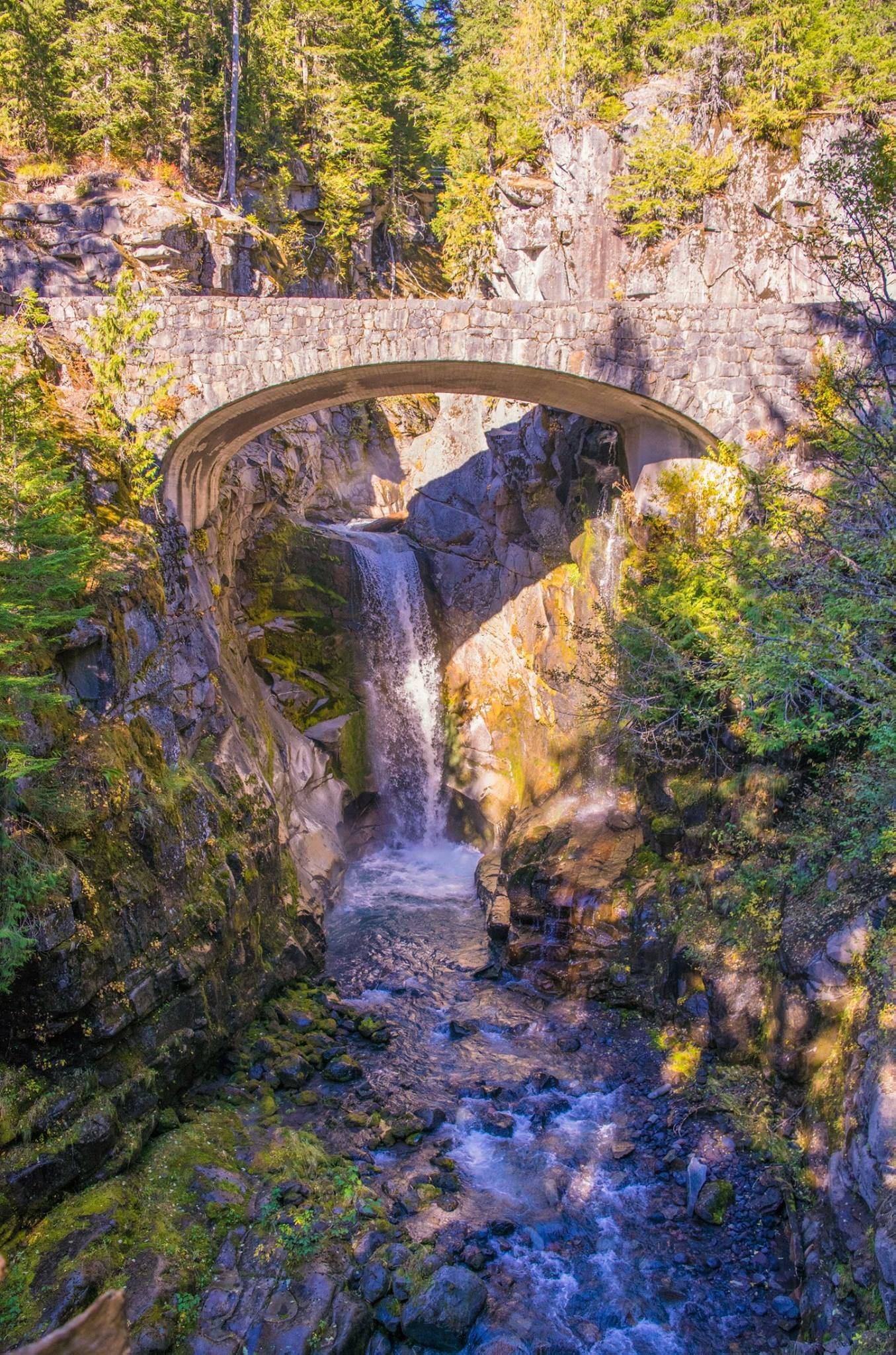Historic stone arch bridge spans over cascading waterfall in dense forest setting.