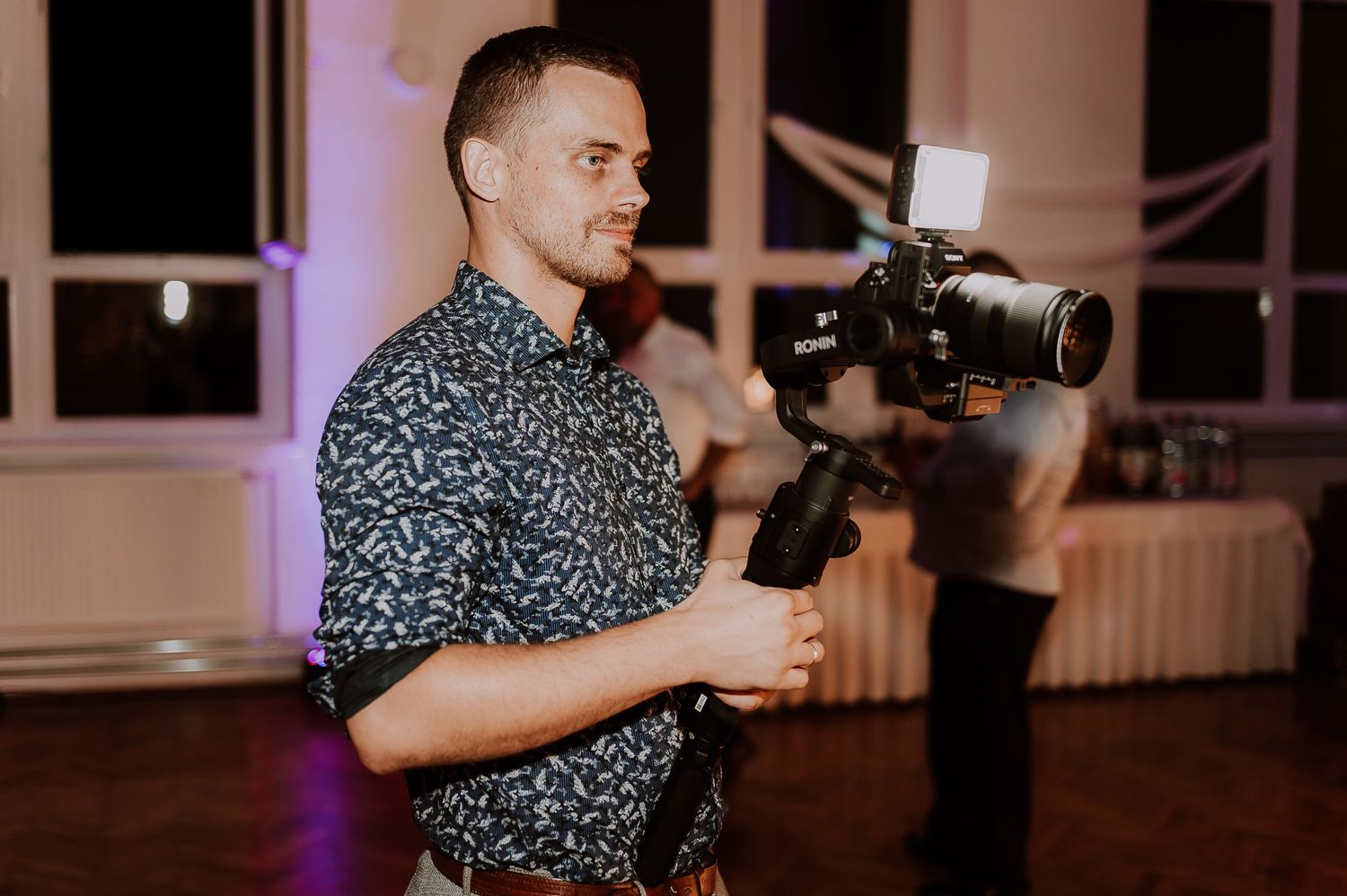 A videographer in a patterned button-down shirt operates a professional camera gimbal stabilizer during an indoor event.