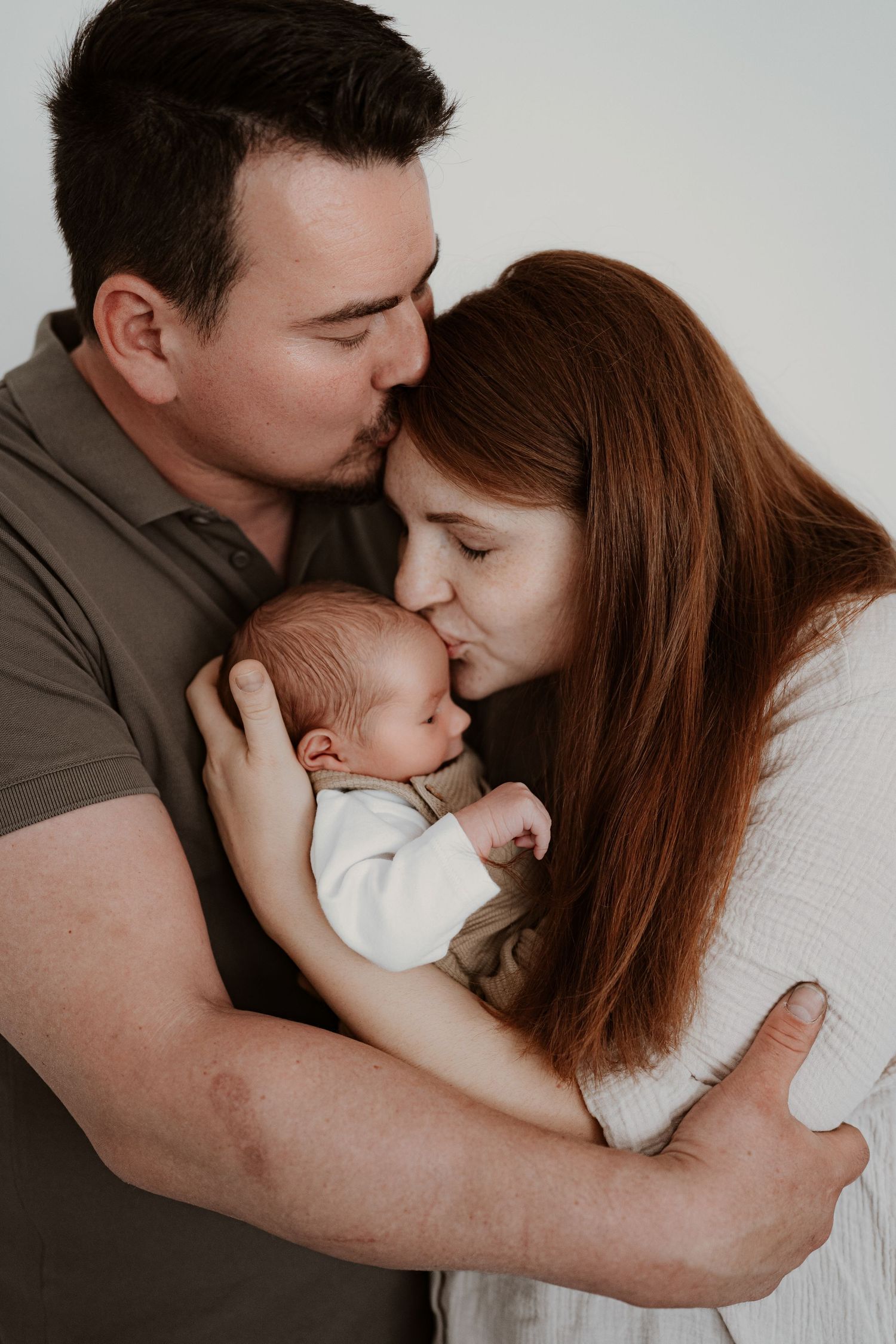 Intimate family portrait shows loving parents embracing and kissing their newborn baby against light background.